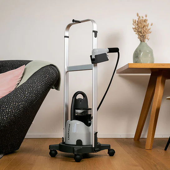 Wheeled garment steamer with a tall metal frame and attached hose, standing on a wooden floor beside a sofa and a table.