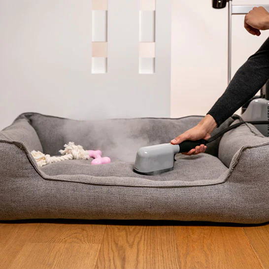 Person uses a handheld steam cleaner on a gray pet bed, steam rising as a blue toy and rope lie nearby on a wooden floor.