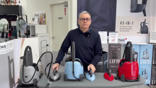 Store clerk stands behind a counter displaying five vacuum cleaners in gray, blue, and red in a showroom setting.
