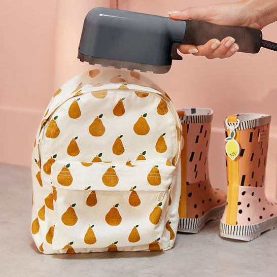 Person uses a handheld garment steamer to press a white backpack with a pear pattern, with pink rain boots nearby.
